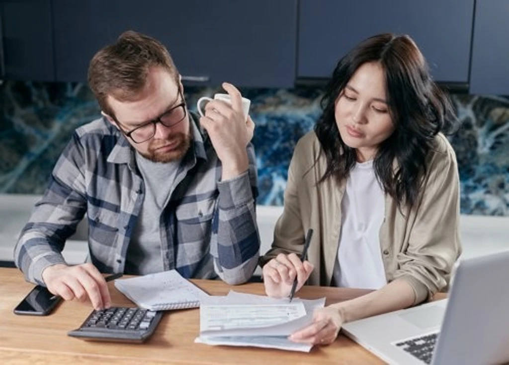 A couple sits at a table reviewing bills with a laptop, calculator, and paperwork